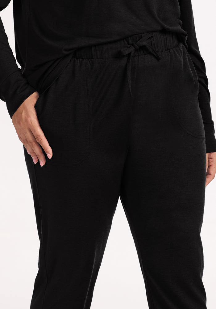A person wears Woolx Mary Kate Lounge Pants in black with a drawstring waist, left hand in pocket. These comfy, moisture-wicking Merino wool pants are perfect for travel, lounging, or relaxing at home. The background is plain and light-colored.