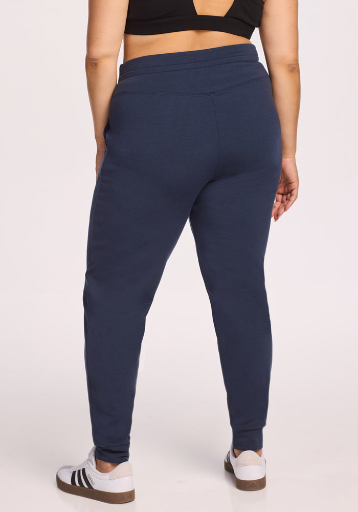 A person wearing Woolx Willa Joggers in Deep Navy, a black crop top, and white sneakers with black stripes stands facing away from the camera against a plain light background.