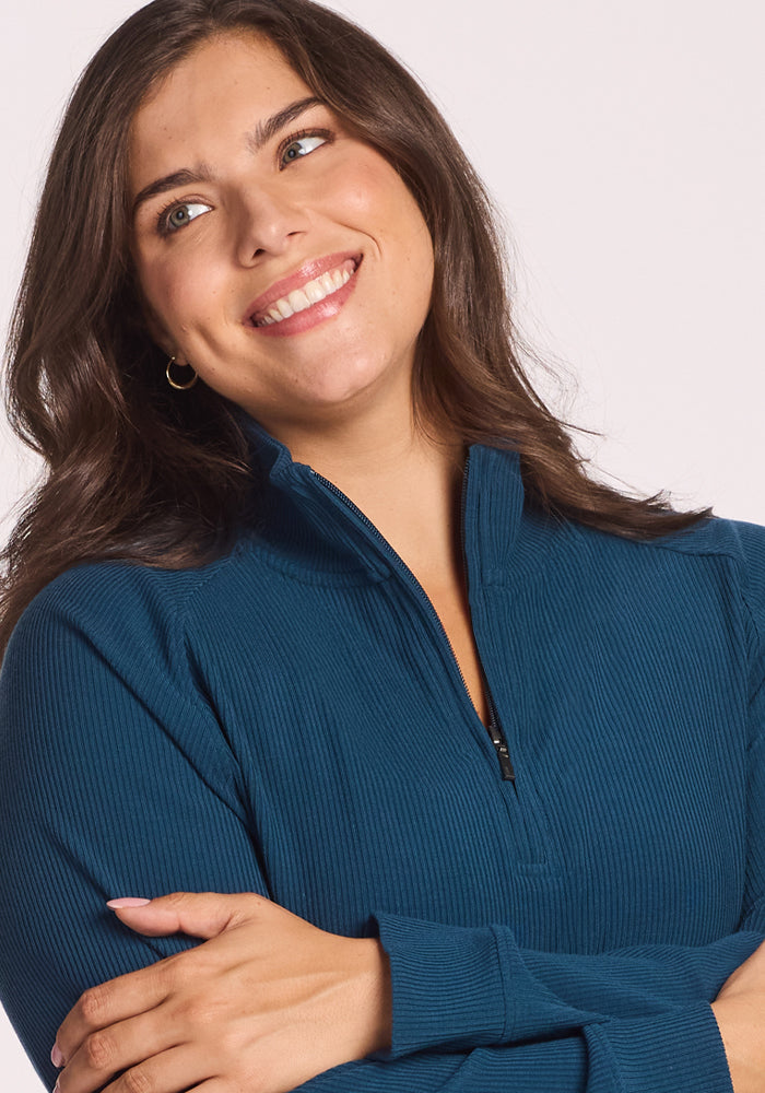 A woman with long brown hair, wearing the Woolx Quinn Ribbed 1/4 Zip in Majolica Blue, smiles and looks to the side with her arms crossed.
