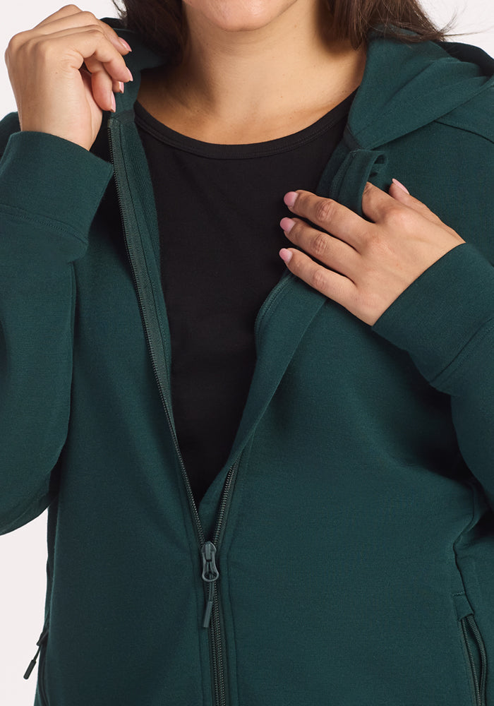 A woman wears the Woolx Darby Zip Up Hoodie in Deep Timber over a black shirt, holding the zipper and hood. The image is cropped to her torso and hands, not showing her full face.