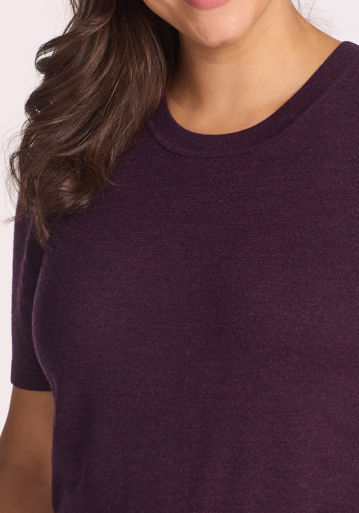 A woman with wavy brown hair is wearing the Woolx Anna Crew Sweater in Deep Plum. Only her mouth, neck, upper chest, and part of her arm are visible as she smiles slightly.