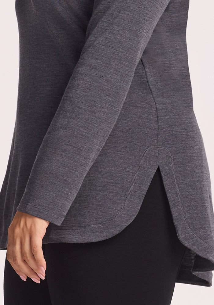 A person wears the Woolx Blair Tunic in Cinder Melange—a long-sleeve Merino wool top with a curved, split hem—over black leggings. The side view highlights the tunic's unique hem and lightweight warmth.