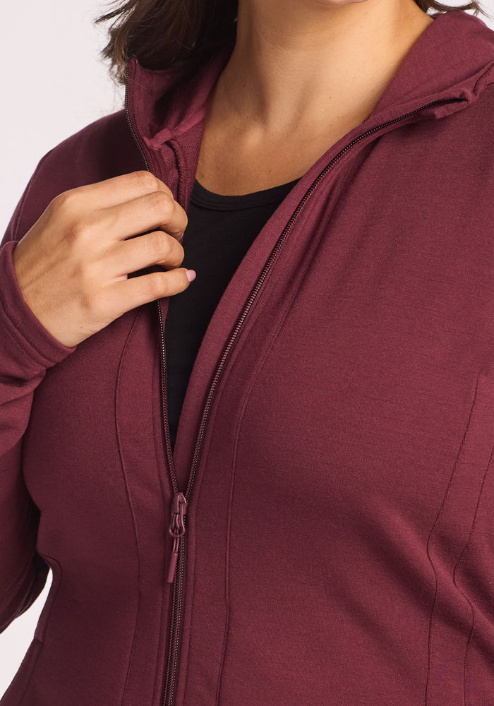 A person wearing a Woolx Zoey Zip Up Hoodie in Toasted Garnet over a black shirt is partially unzipping the merino wool hoodie, with their hand holding the zipper near the collar.