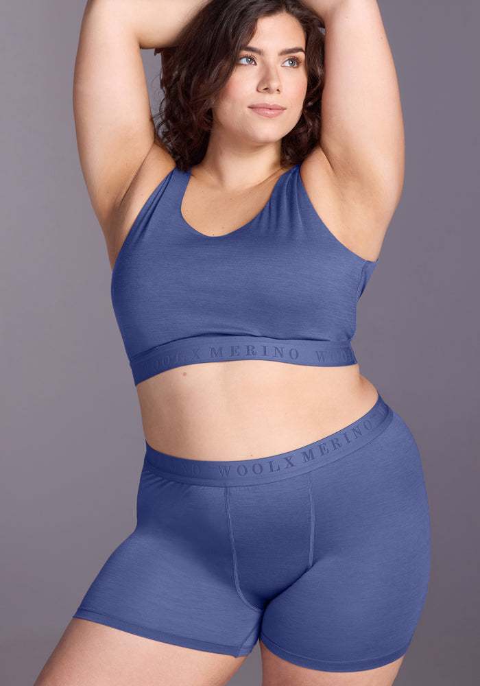 A woman with curly brown hair poses confidently in a matching blue sports bra and Woolx Elliott Boyshort - Cloudy Bay set, featuring "WOOLX MERINO" on the waistband, against a plain background.