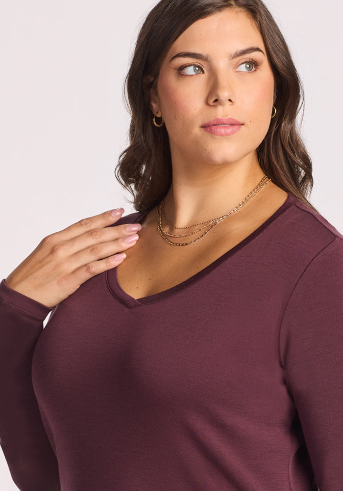 A woman with wavy brown hair, light makeup, and layered gold necklaces wears the Woolx Eva Tunic in Chocolate Fig. She looks to her left, gently touching her collar. The background is plain and light-colored.