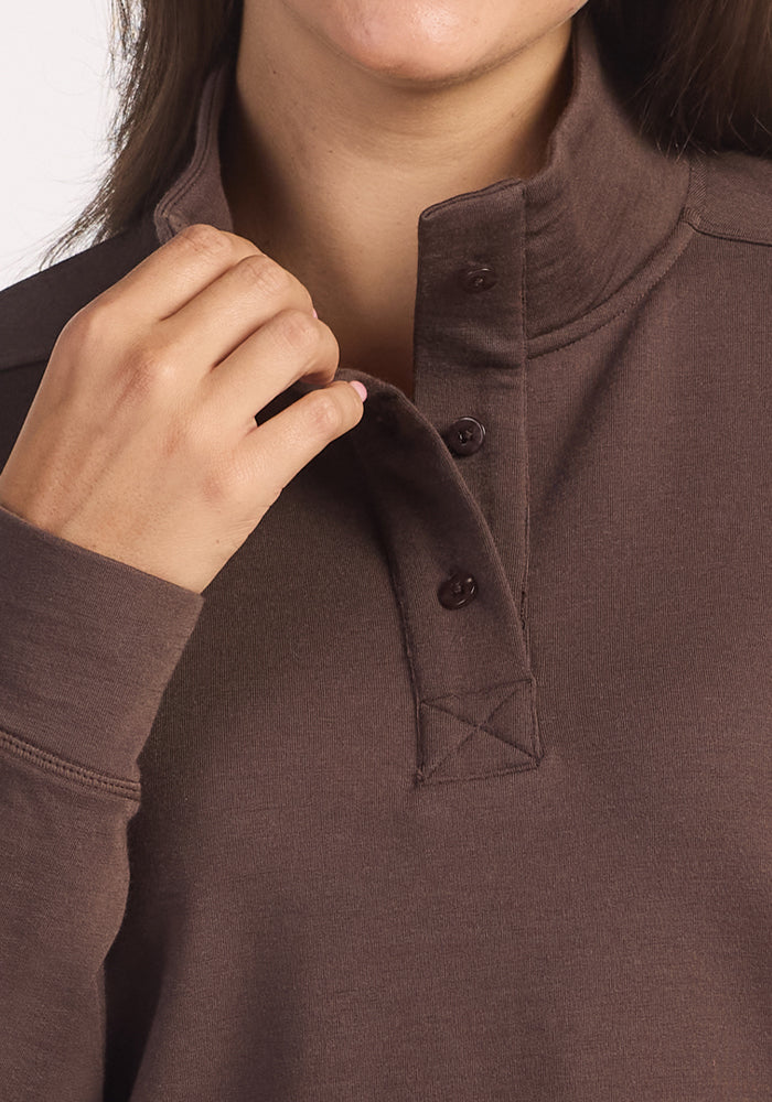 Close-up of a person wearing the Woolx Annie 3 Button Pullover in Bracken, touching the collar with their left hand; only the lower part of their face is visible.