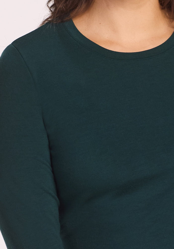 A close-up of a person wearing the Woolx Sadie Baselayer Top in Deep Timber, made from Duralite merino fabric, showing their chest, shoulder, and neck with wavy brown hair partially visible.