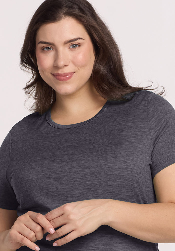 A woman with long brown hair smiles gently at the camera, wearing the Woolx Liza Crew Neck Tee in Cinder Melange, a short-sleeved lightweight shirt, and stands against a light background with her hands together in front of her.