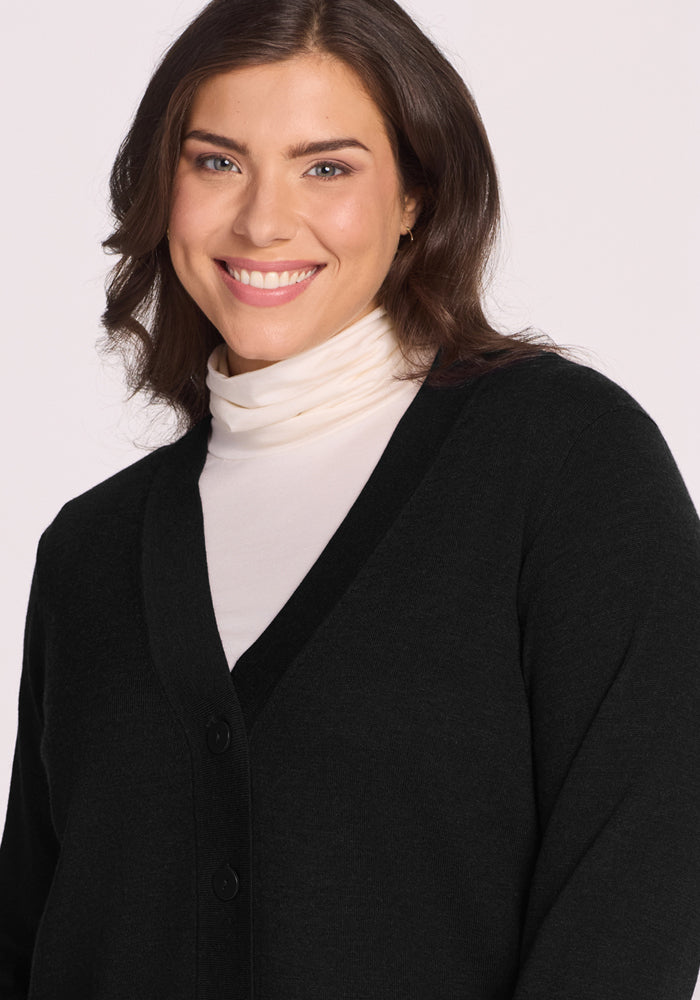 A woman with wavy brown hair, wearing the Woolx Gia Sweater in black and a white turtleneck, smiles at the camera against a light background.