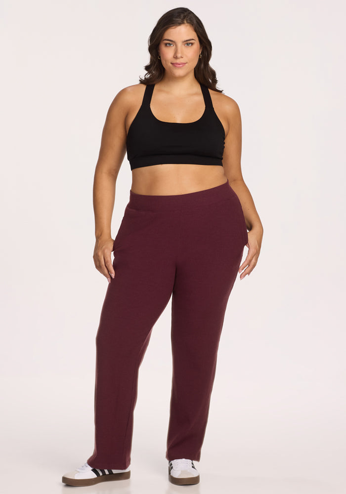 A woman with long brown hair wears a black sports bra and Woolx Luca Ribbed Pants in Toasted Garnet, paired with white sneakers with black stripes. She faces forward against a plain white background, smiling slightly.