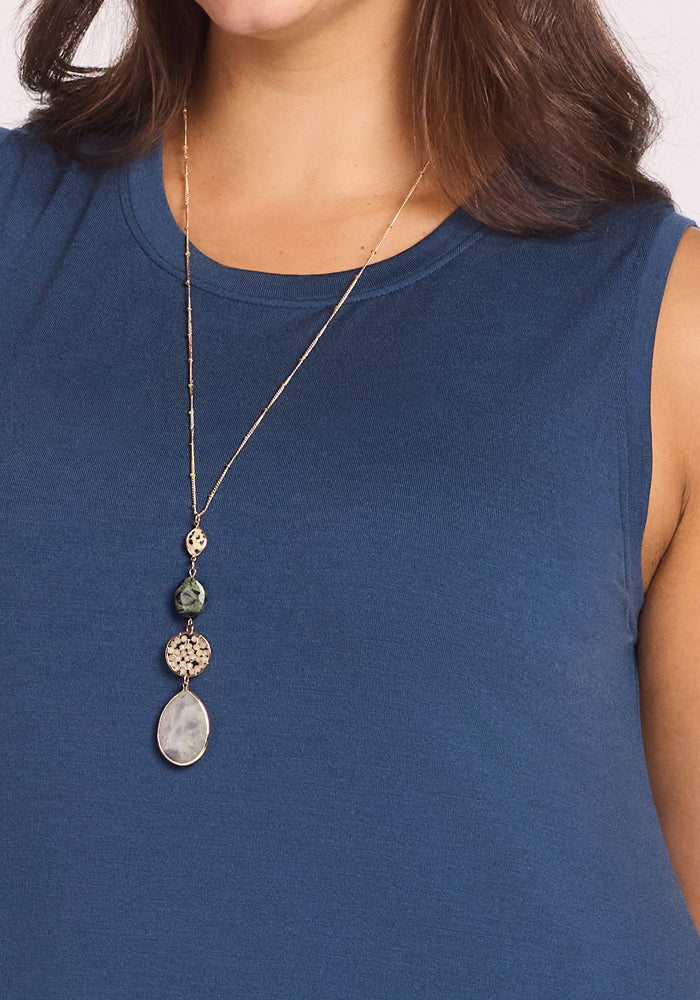 A woman wearing the Woolx Clara Dress in Starry Night, a sleeveless blue breathable summer dress, paired with a long gold necklace featuring decorative beads and a large teardrop-shaped pendant.