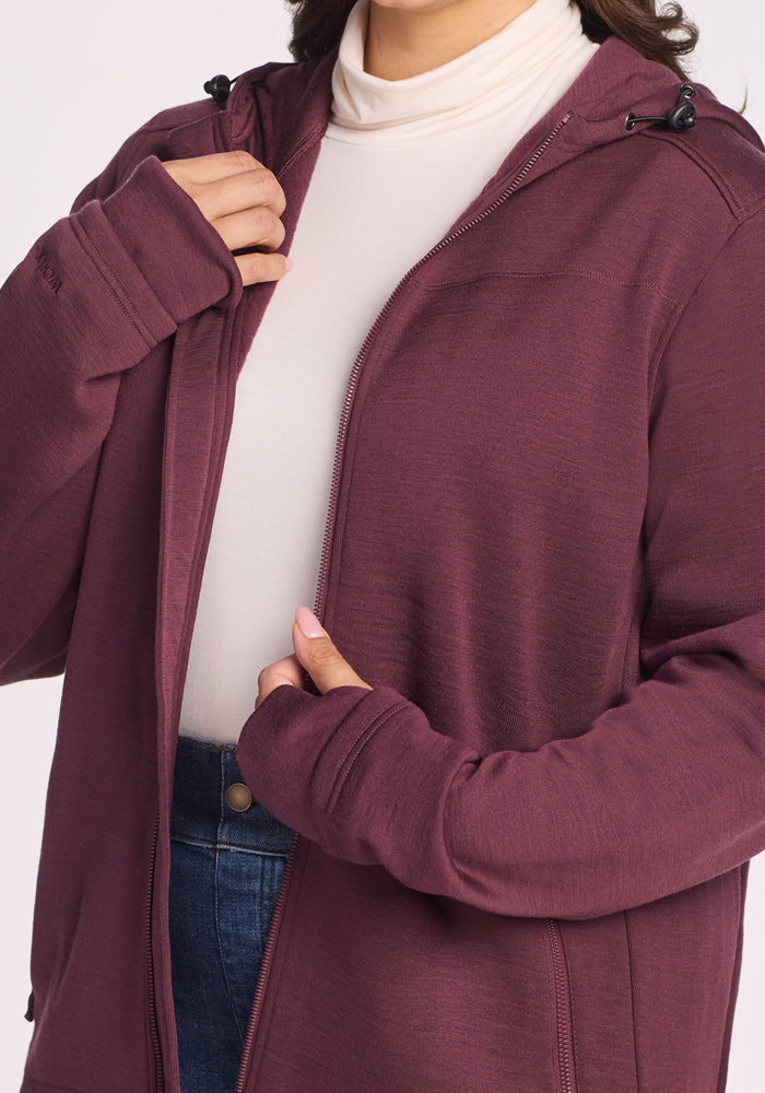 A person wears the Woolx Cubby Hooded Sweatshirt in Grape Harvest over a white turtleneck and blue jeans, holding the open hoodie near the chest. Their face is partially out of frame.