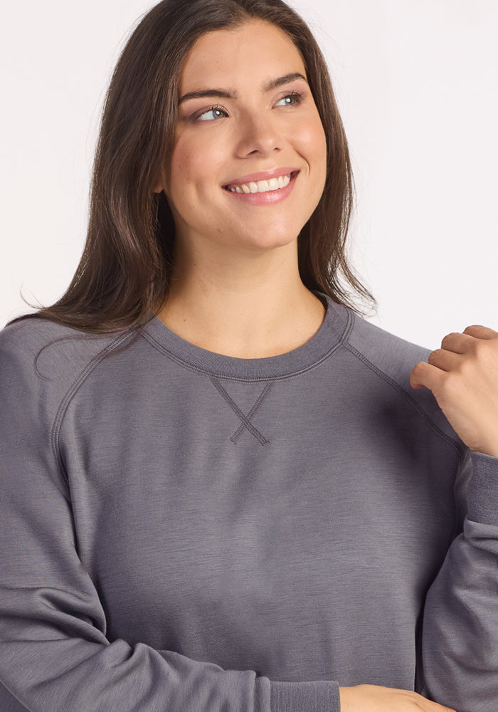 A woman with long brown hair smiles and looks to the side as she poses in the Woolx Bailey Crewneck Sweatshirt - Arctic Dusk, enjoying its cozy Merino wool warmth and breathable comfort in a classic crewneck style.