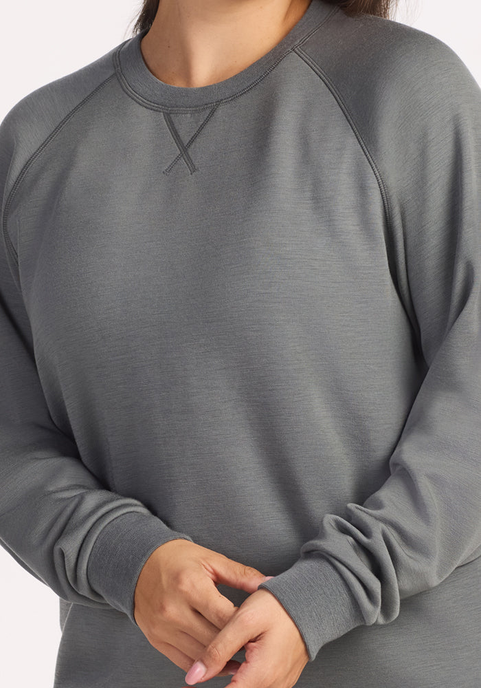A person wears the Woolx Bailey Crewneck Sweatshirt in Sedona Sage, featuring long sleeves, a subtle X-stitch below the collar, and made from recycled polyester; their hands are gently clasped, face partially out of frame.