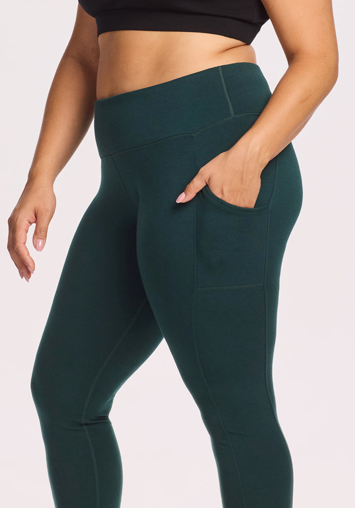Someone wearing Woolx Piper Pocket Leggings in Deep Timber, featuring a high waist, moisture-wicking fabric, a side pocket, and paired with a black crop top stands against a plain light background.
