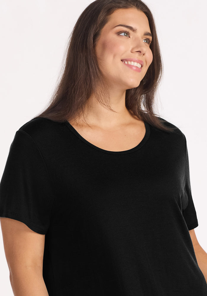 A woman with long brown hair, wearing the Woolx Desi Nightgown in black, smiles and looks slightly to the side against a light background.