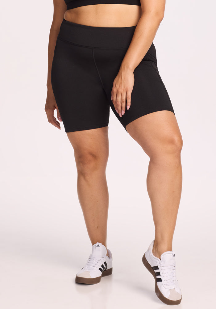 A person wearing a black sports bra, Woolx Merino Flex® Dani Bike Shorts in black, and white Adidas sneakers with black stripes stands with one hand on their thigh against a plain light background.