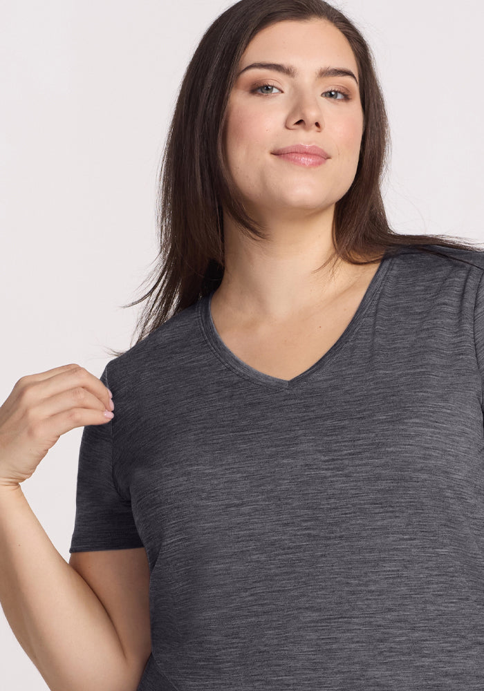 A woman with long brown hair and fair skin wears the Woolx Junie V Neck in Cinder Melange, a relaxed fit women's tee. She faces forward, slightly smiling with one hand near her shoulder, against a plain light background.
