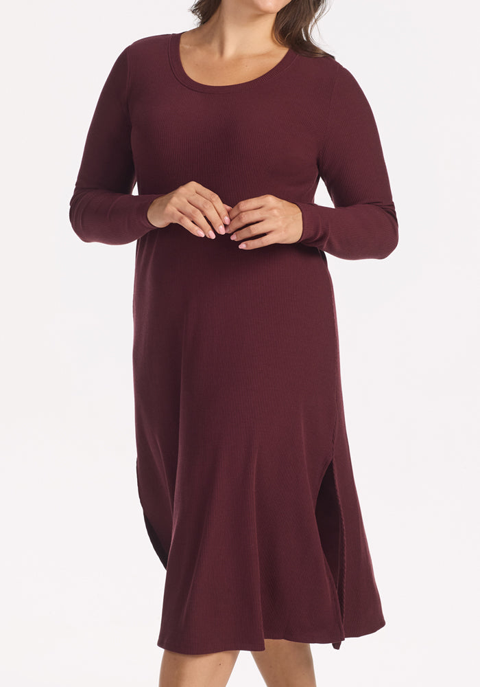 A woman models the Woolx Caroline Ribbed Twirl Dress in Toasted Garnet, a wrinkle-resistant, long-sleeved merino dress with side slits and a knee-length, versatile silhouette. She stands against a white background with her hands clasped, face unseen.