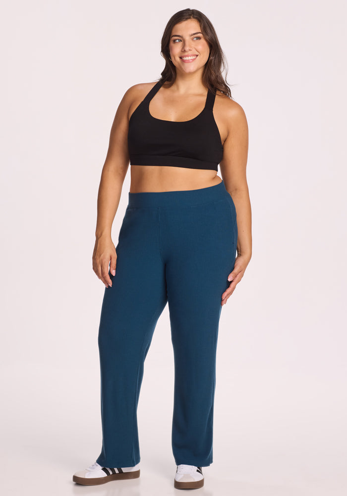 A woman with long brown hair smiles while wearing Woolx's Luca Ribbed Pants in Majolica Blue, a black sports bra, and white sneakers with black stripes, standing against a plain light background.
