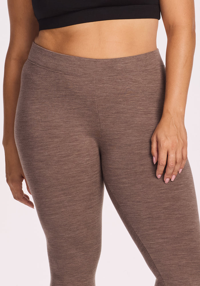 A person is shown from mid-torso to thigh wearing Woolx Stella Leggings in Simply Taupe and a black crop top, with one hand on their hip against a plain background.