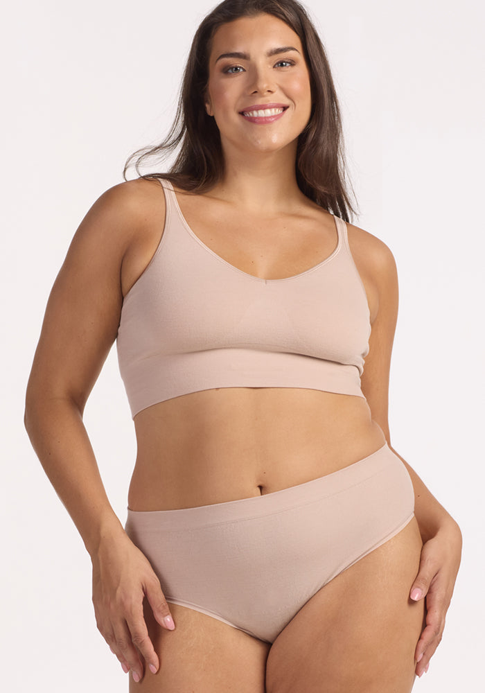 A smiling woman with long brown hair wears the Woolx Maeve Bikini in Sand, standing against a plain light background.