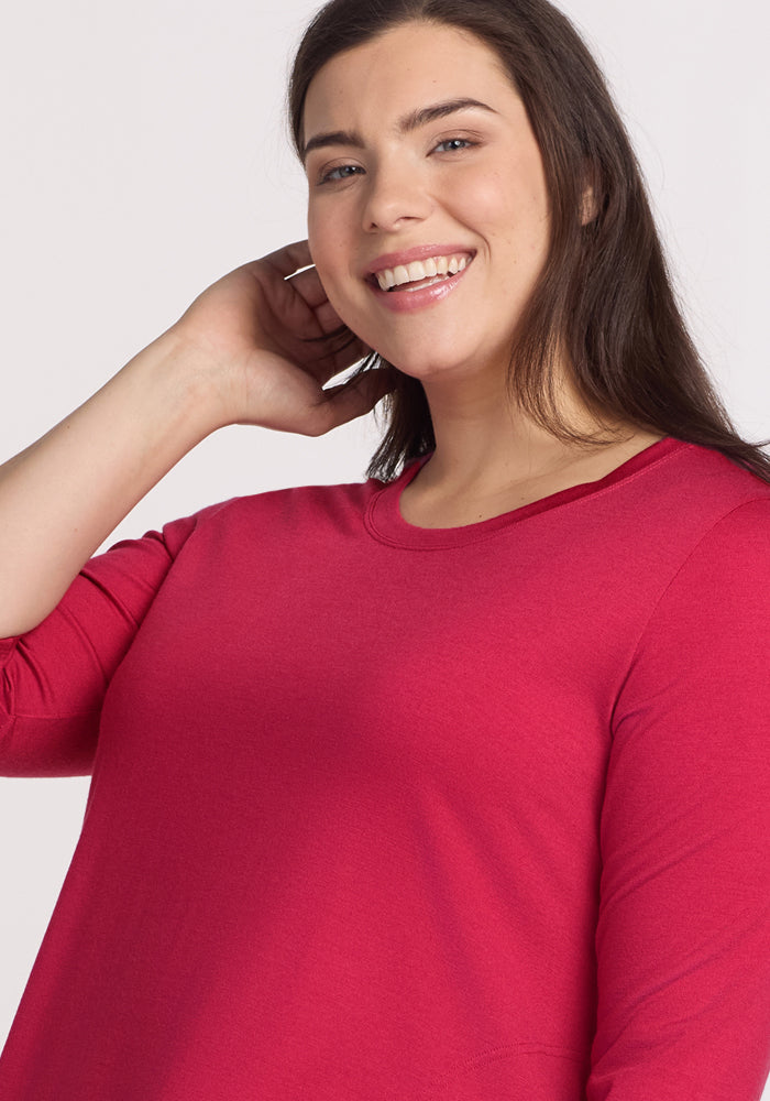 A woman with long brown hair wears the Woolx Rissa A Line Dress in Poppy Spark, smiling at the camera and touching her neck with one hand against a plain white background—perfect for fall and winter layering.