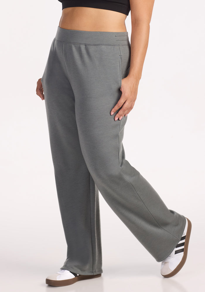 A person wearing Woolx Bree Lounge Pants in Sedona Sage, paired with a black crop top and white sneakers with black stripes, stands with one leg slightly forward against a plain white background.