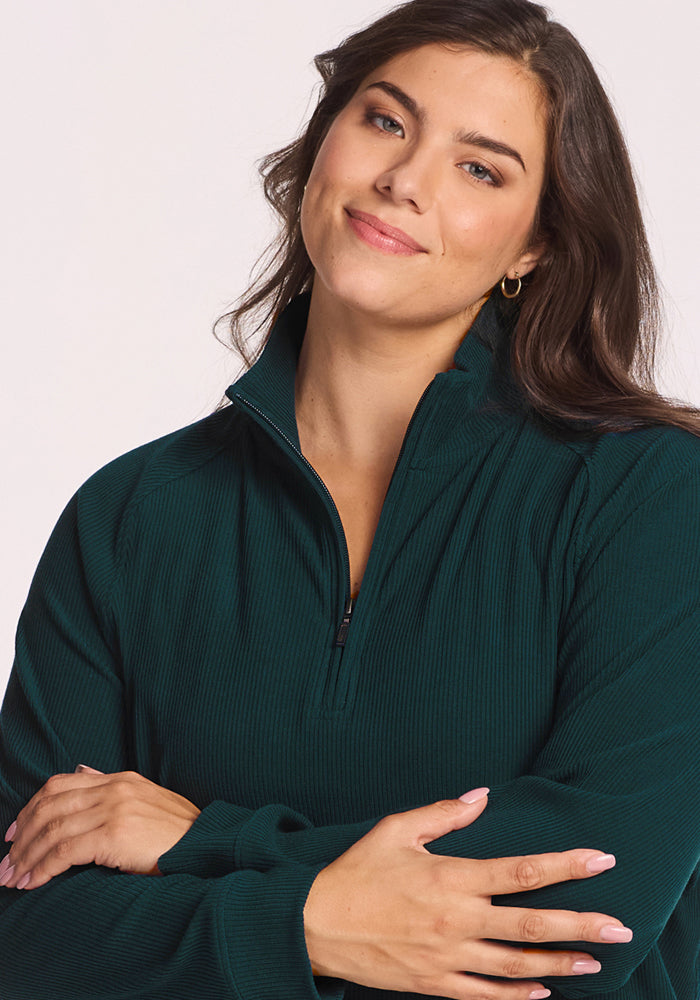 A woman with long brown hair, wearing a Woolx Quinn Ribbed 1/4 Zip in Deep Timber, smiles softly with her arms crossed in front of her against a light background.