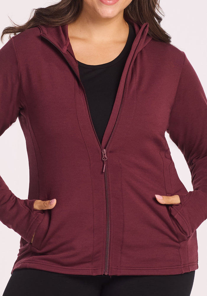 Someone wearing a Woolx Zoey Zip Up Hoodie in Toasted Garnet, partially unzipped over a black top, with hands in the front pockets. The hoodie is shown from shoulders to hips for a relaxed, casual style.