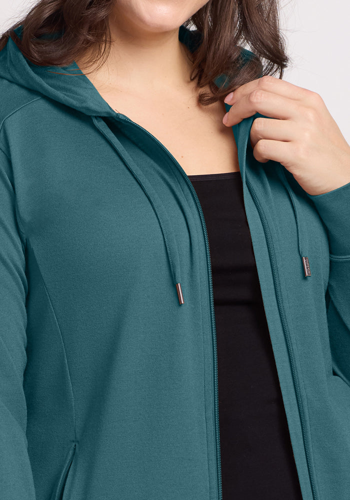 A person wearing the Woolx Ryann Hoodie in Tidepool over a black top holds the hood’s edge, with their upper torso and part of their face visible, highlighting this essential travel piece.