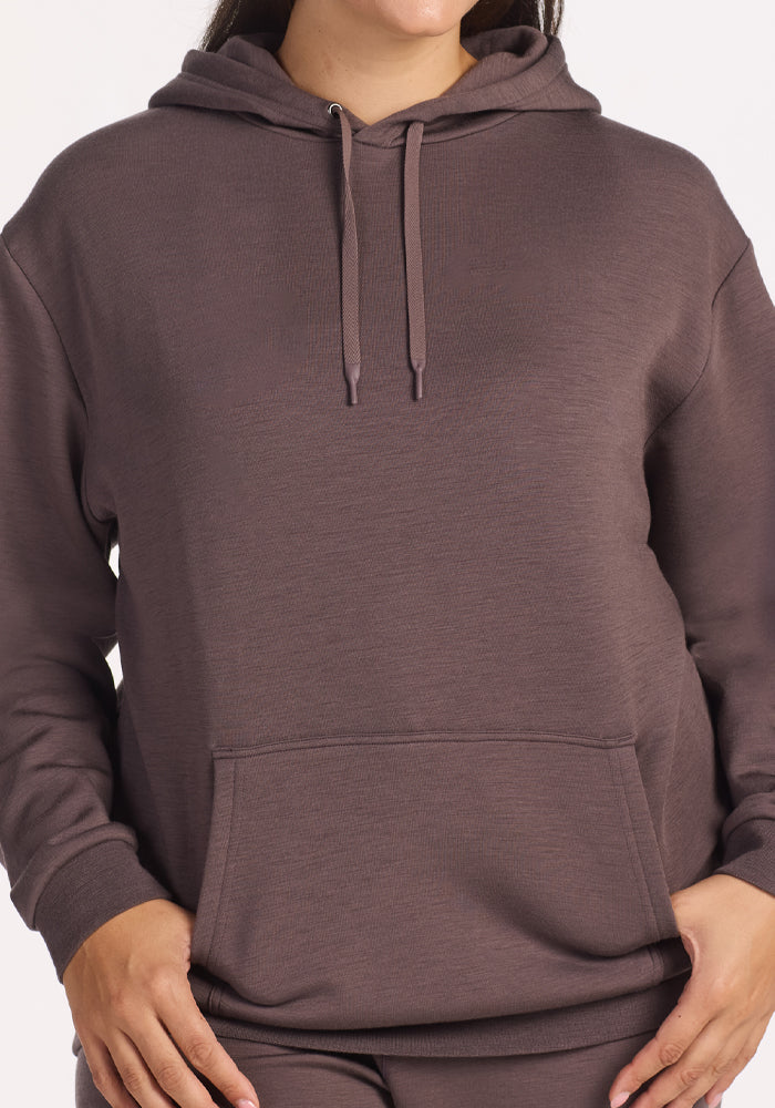 A person wears the Woolx Avery Hoodie in Plum Truffle, featuring recycled polyester, a front pocket, drawstrings, and long sleeves. The image is cropped at the shoulders and thighs, showing only part of the face.