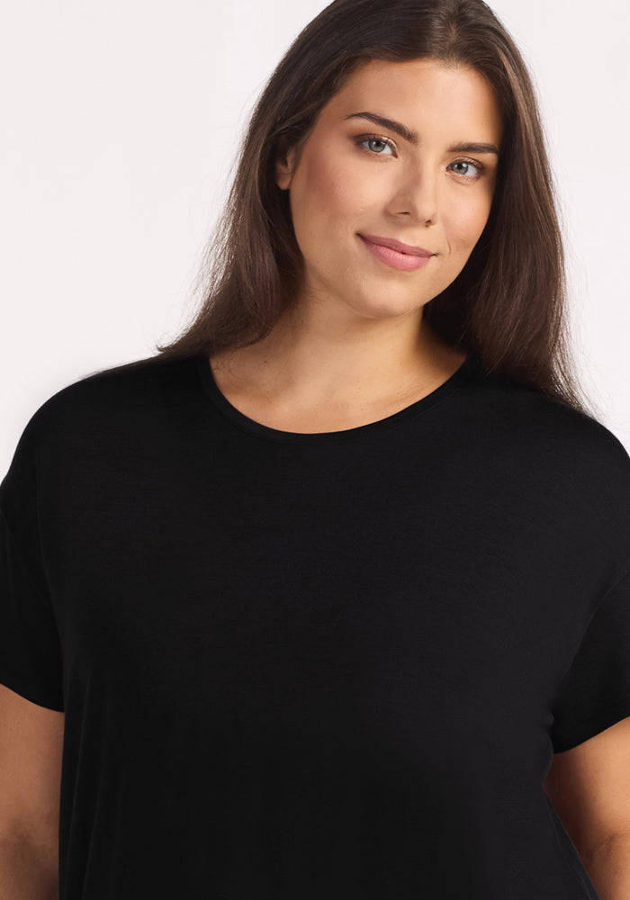 A woman with long brown hair and light skin smiles softly, wearing the Woolx Billie Lounge Tee in black, standing against a simple white background.