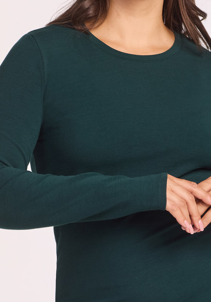 A woman wears the Woolx Sadie Baselayer Top in Deep Timber, a long-sleeve dark green thermal, with her hands gently clasped in front, shown from shoulders to waist.