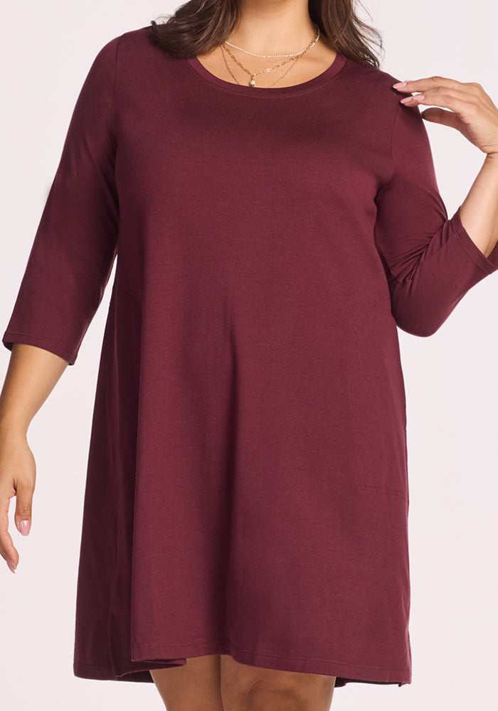 A woman models the Woolx Rissa A Line Dress in Toasted Garnet, a maroon, knee-length Merino wool dress with long sleeves and a round neckline. Perfect for fall and winter layering, it offers simple, casual style for travel.