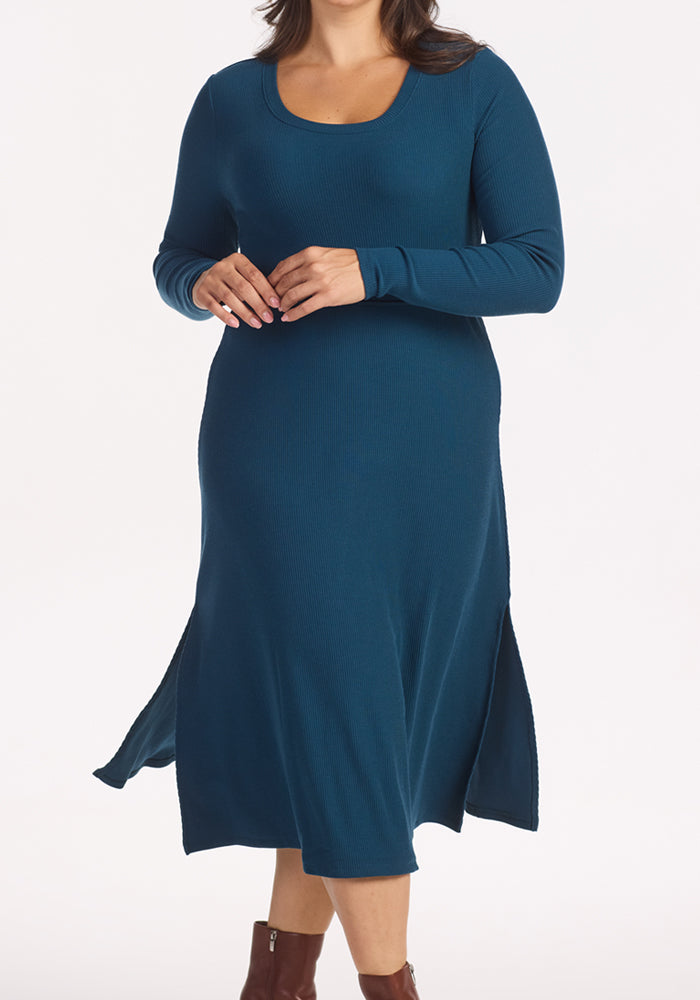 A person wearing the Woolx Caroline Ribbed Twirl Dress in Majolica Blue, featuring long sleeves, side slits, and a scoop neckline, stands against a white background with partially visible brown ankle boots.