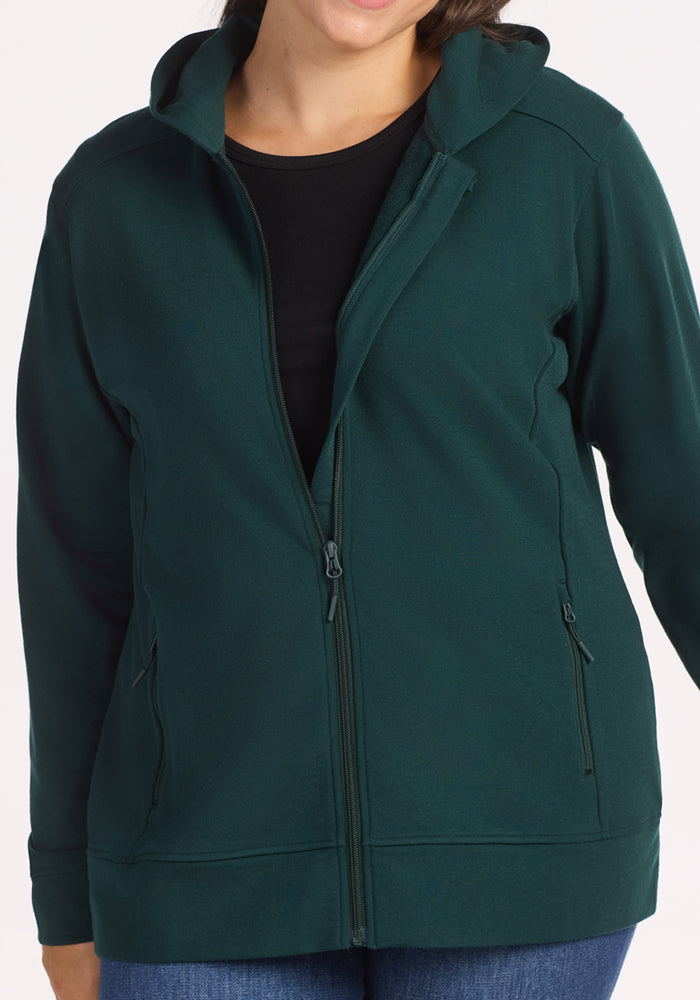 A woman wearing the Woolx Darby Zip Up Hoodie in Deep Timber, made from Duralite merino, over a black shirt and blue jeans stands with arms relaxed. The hoodie features zippered pockets and a hood. Her face is partially out of frame.