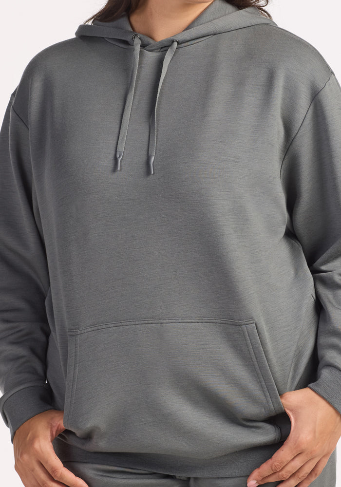 A person wears the Woolx Avery Hoodie in Sedona Sage, a loose-fitting antimicrobial hoodie with drawstrings and a front pocket, hands partially in the pocket. The cropped image shows only the body and hoodie sleeves.