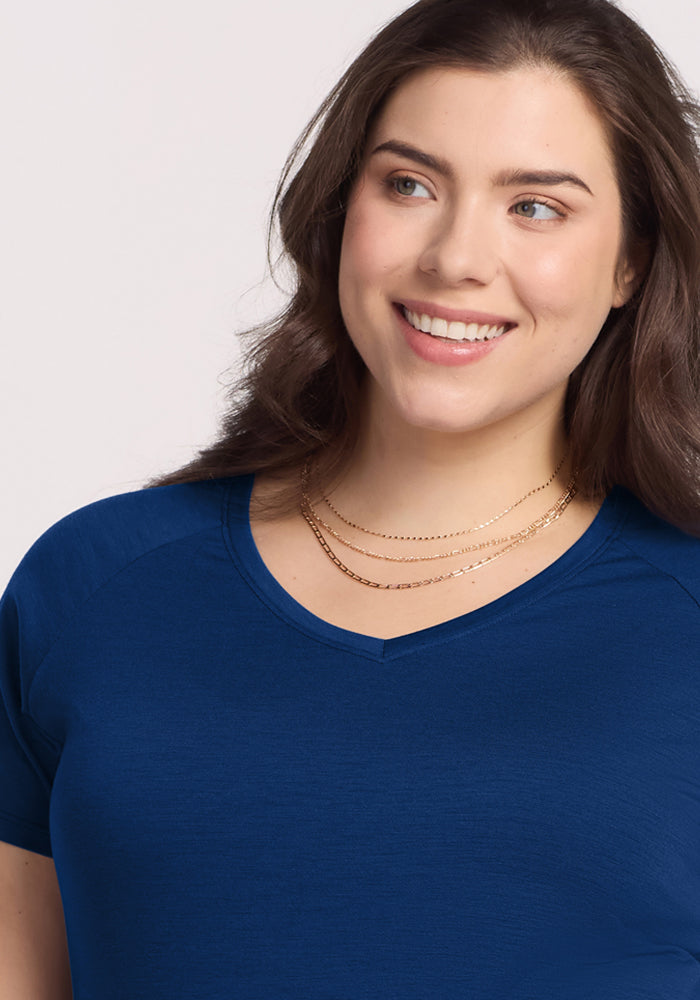 A woman with long brown hair, wearing the Woolx Mia V Neck in Estate Blue and layered gold necklaces, smiles as she looks slightly to the side.