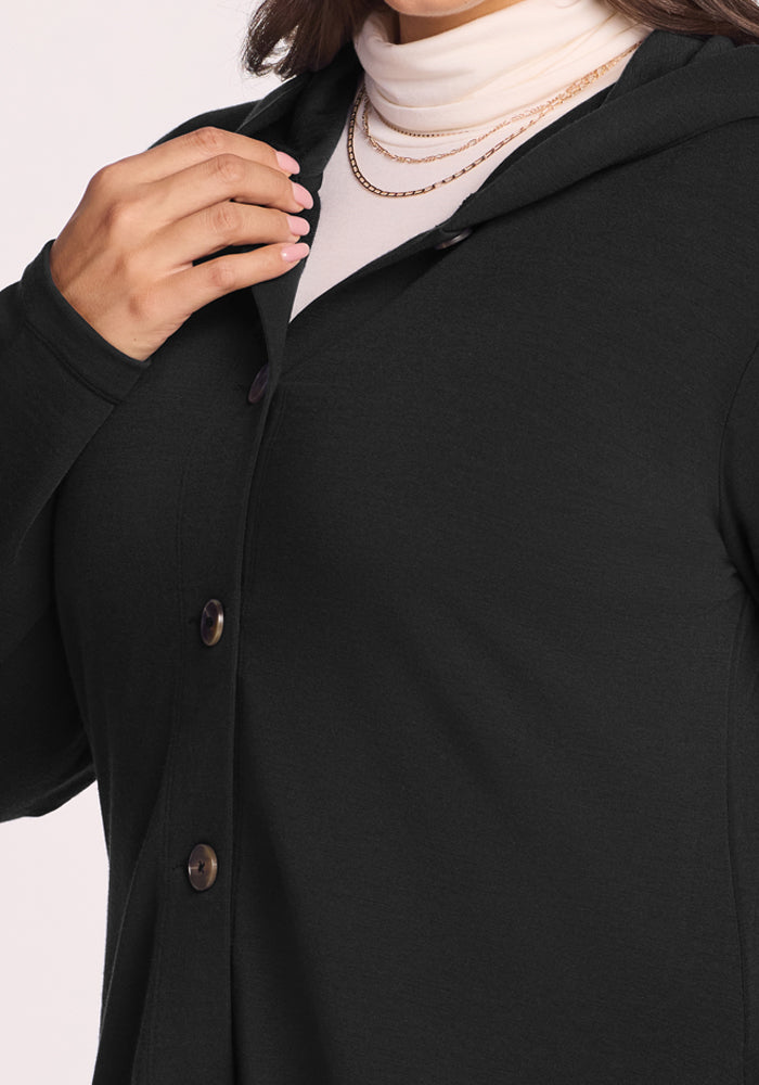 A person wearing the Woolx Edna Hooded Cardigan in black over a white turtleneck, layered with gold necklaces, gently holds the cardigan's collar.