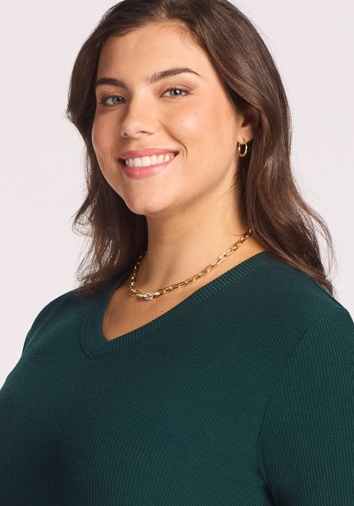 A woman with long brown hair wears the Woolx Evie Ribbed Tunic in Deep Timber, paired with gold hoop earrings and a gold chain necklace. She smiles and looks slightly to the side against a plain light background.