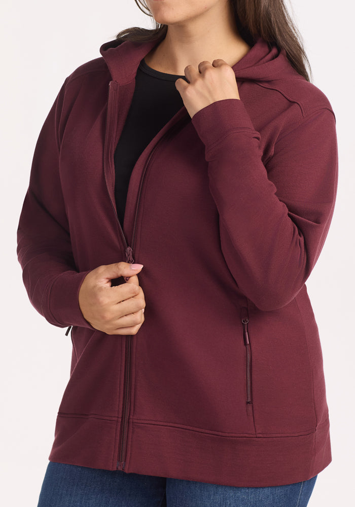 A woman wears the Woolx Darby Zip Up Hoodie in Toasted Garnet over a black shirt and blue jeans, standing against a light backdrop with one hand near the collar and the other by the zipper.