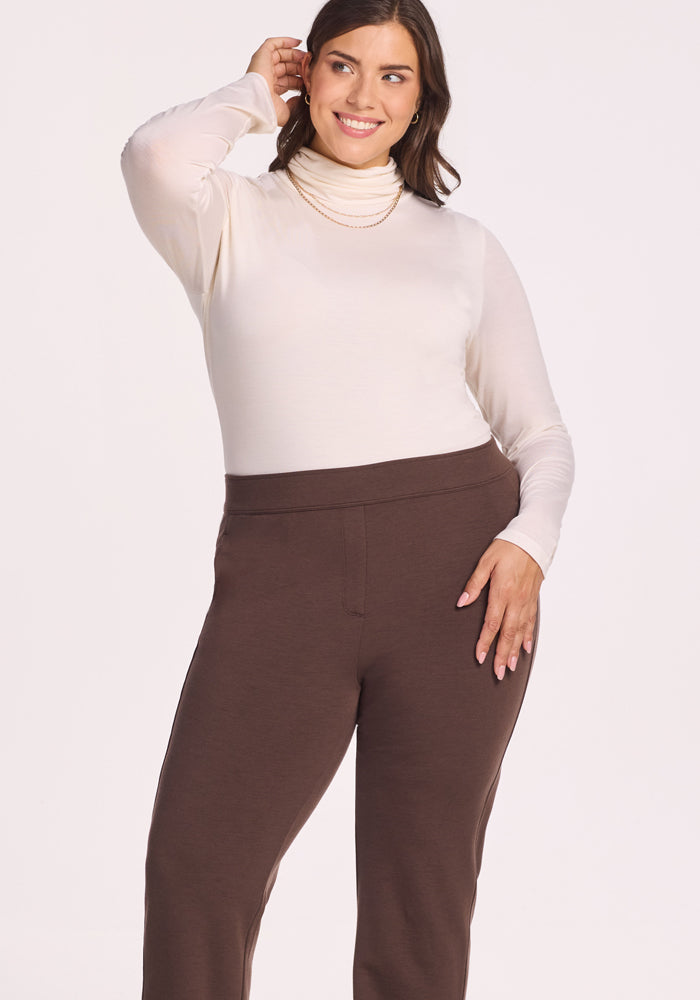 A woman smiles while touching her hair, wearing a long-sleeve white turtleneck top and Woolx Emerson Straight Leg Pants in Bracken against a plain light background.