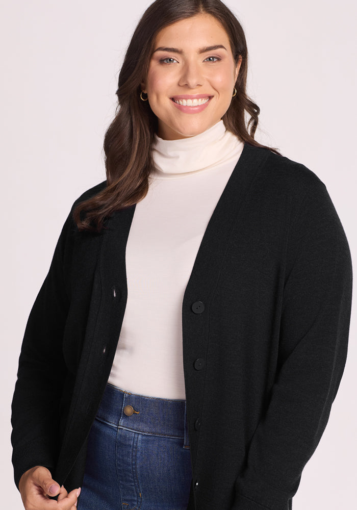 A woman with long brown hair smiles in a white turtleneck, the Woolx Gia Sweater - Black cardigan, and blue high-waisted jeans against a light-colored background.