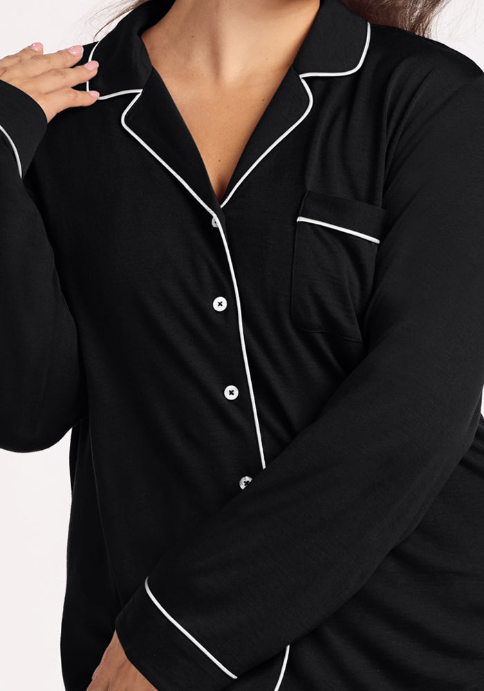 A person wears the Woolx Harper FeatherTouch® Pajama Set in black, featuring white piping, white buttons, and a chest pocket. Their hand is raised near the collar against a plain, light-colored background.