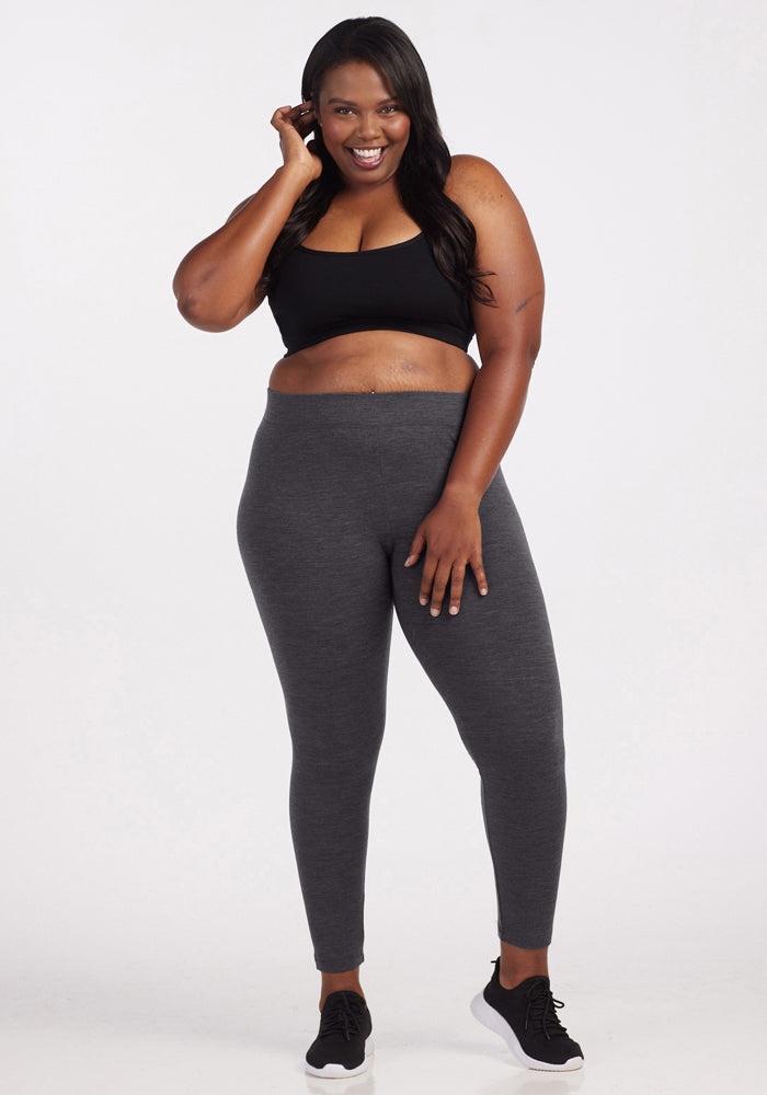 A smiling woman with long dark hair confidently models Woolx's Stella Leggings in Cinder Melange, styled with a black crop top and sneakers against a plain white background.