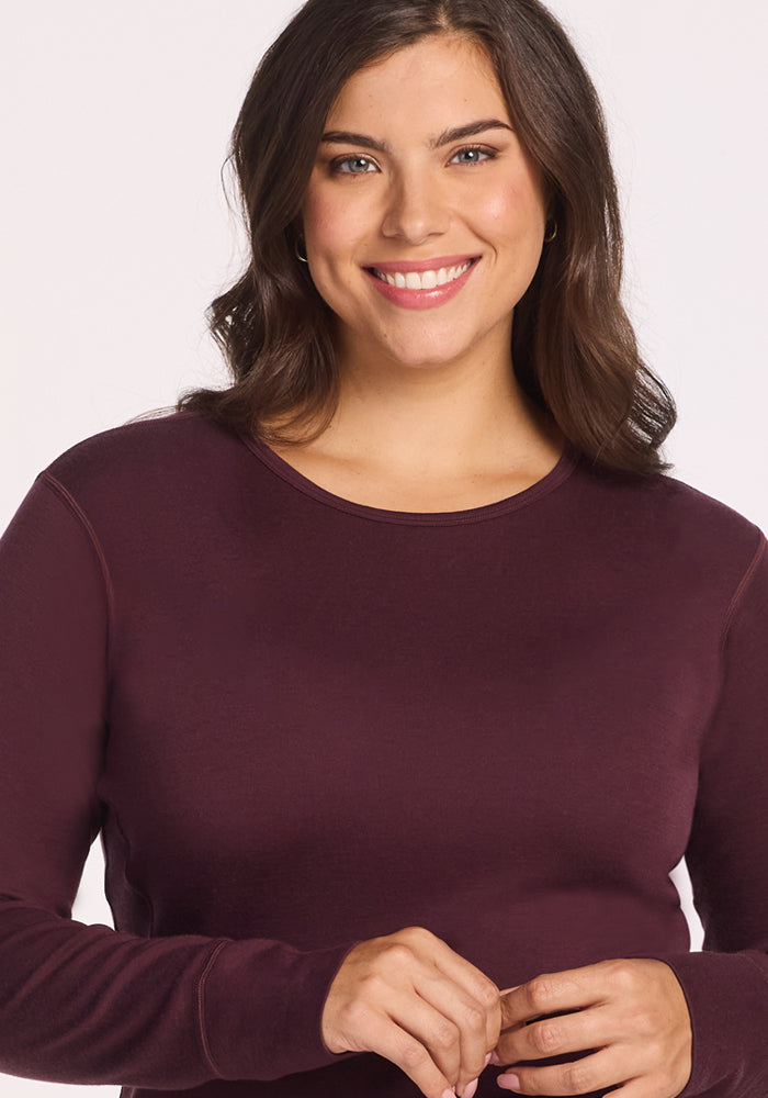 A woman with wavy brown hair is smiling and wearing the Woolx Hannah Long Sleeve Top in Chocolate Fig, standing against a plain, light background.