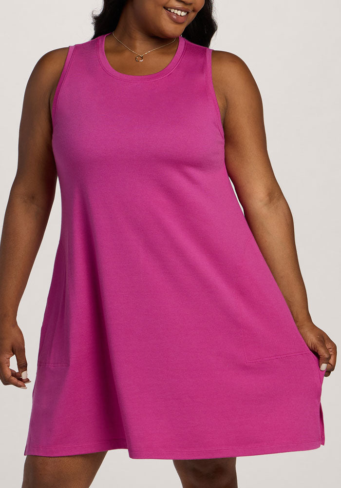 A woman smiles, holding the hem of the Woolx Clara Dress - Summer Sorbet, a bright pink, lightweight style with pockets.