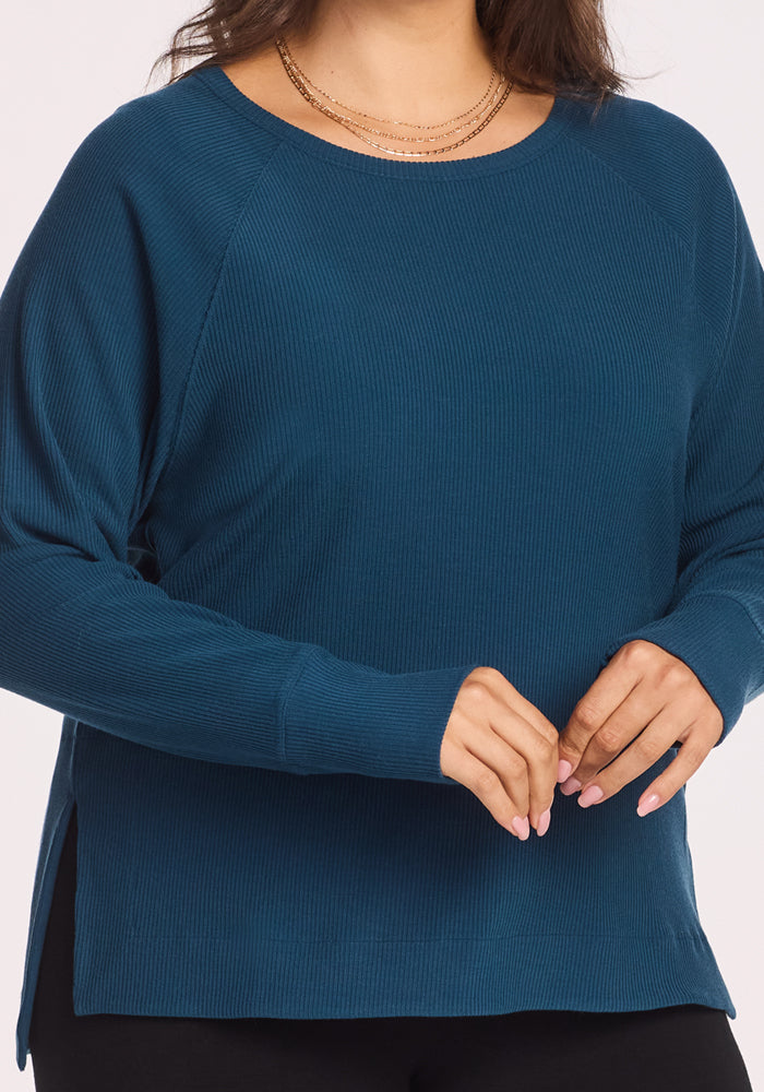 A woman wearing the Woolx Ainsley Sweater in Majolica Blue, paired with layered gold necklaces, stands with her hands gently clasped. Only her torso appears against a plain white background.