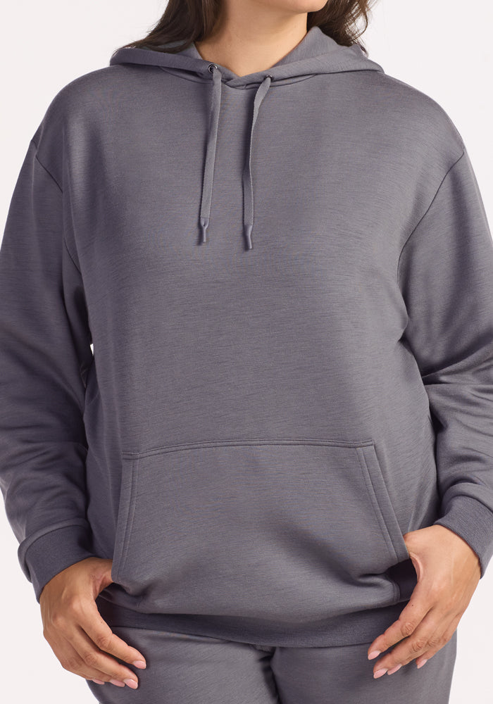 A woman, visible from the torso up, wears the Woolx Avery Hoodie in Arctic Dusk—a loose-fitting, gray antimicrobial hoodie with drawstrings and a front pocket, her hands resting near the pocket.